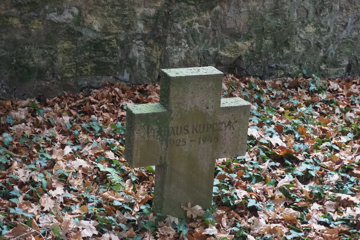Kamienny krzyż porośnięty mchem, wśród liści.
Napis: „?ADÄUS KUP?ZYK 1925–1946”
