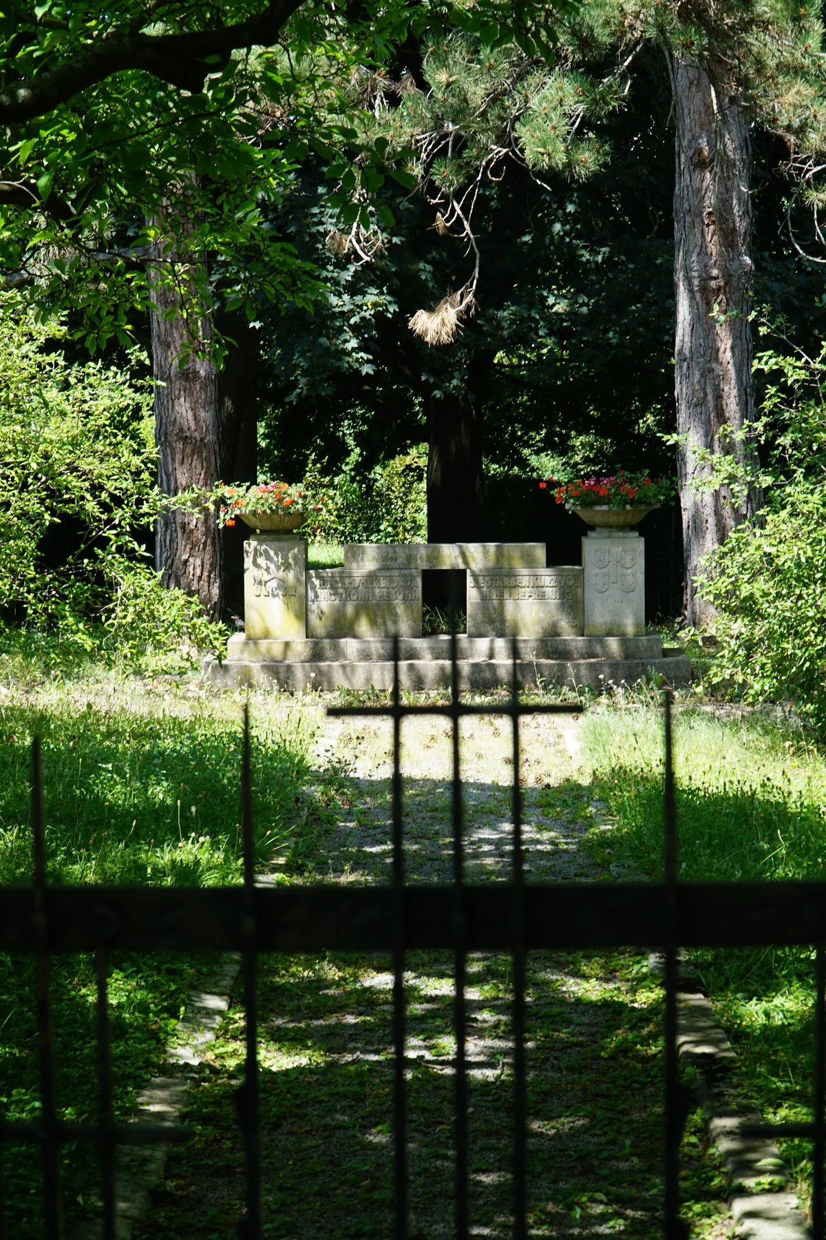 Widok pomnika przez kratę bramy cmentarza; aleja prowadząca do pomnika wśród drzew.