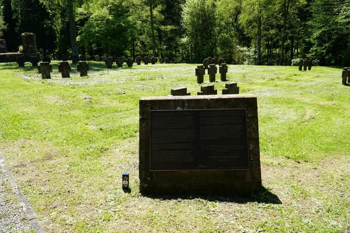 Widok cmentarza KZ: na pierwszym planie kamienny blok z tablicą z brązu, w tle grupy kamiennych krzyży na trawie.