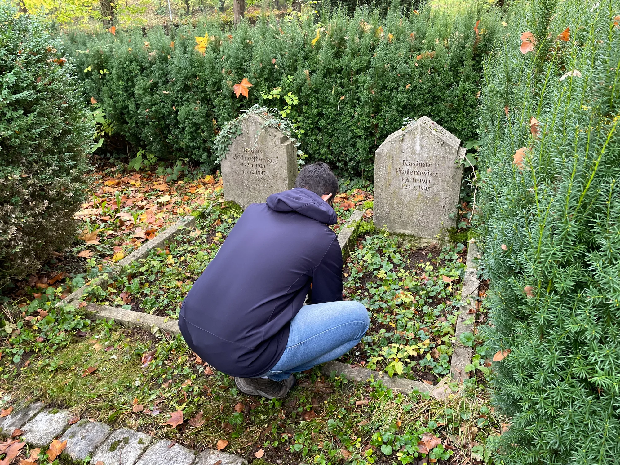 Dwa kamienne nagrobki z obramowaniem, mężczyzna (od tyłu) przy pracach porządkowych.
Napis: „Tadeus Jedrzejewski 23.4.1924 3.12.1947 | Kasimir Walerowicz *6.11.1911 †2.3.1945”